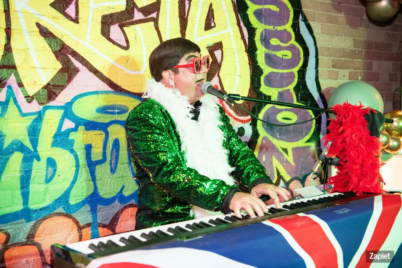 The Elton John tribute performer, in a sequined green jacket, feather boa, and star-shaped sunglasses, plays piano and sings in front of a colorful graffiti-style mural at Zapiet's YOLO Party.