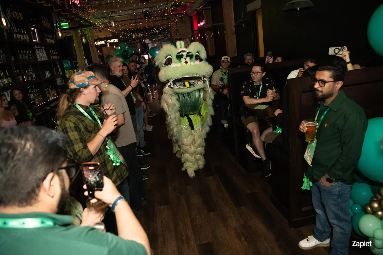 A traditional lion dance performer in a green and gold costume moves through the crowd at Zapiet's YOLO Party, with guests taking photos on their phones as it passes.