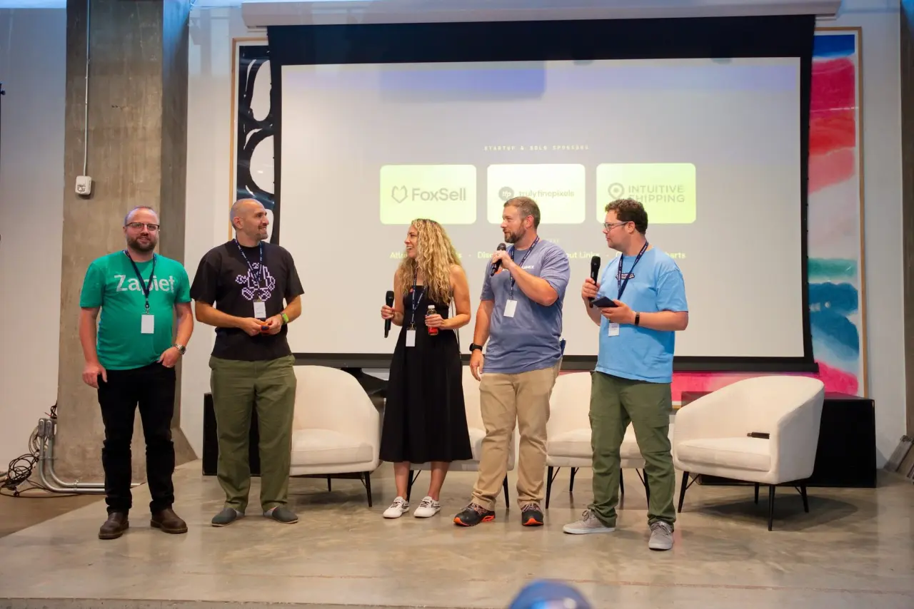 Sam Forde, wearing a green Zapiet t-shirt, stands on stage with four other speakers during the panel at Brightwork Vol.01, with sponsor logos displayed on the screen behind them.