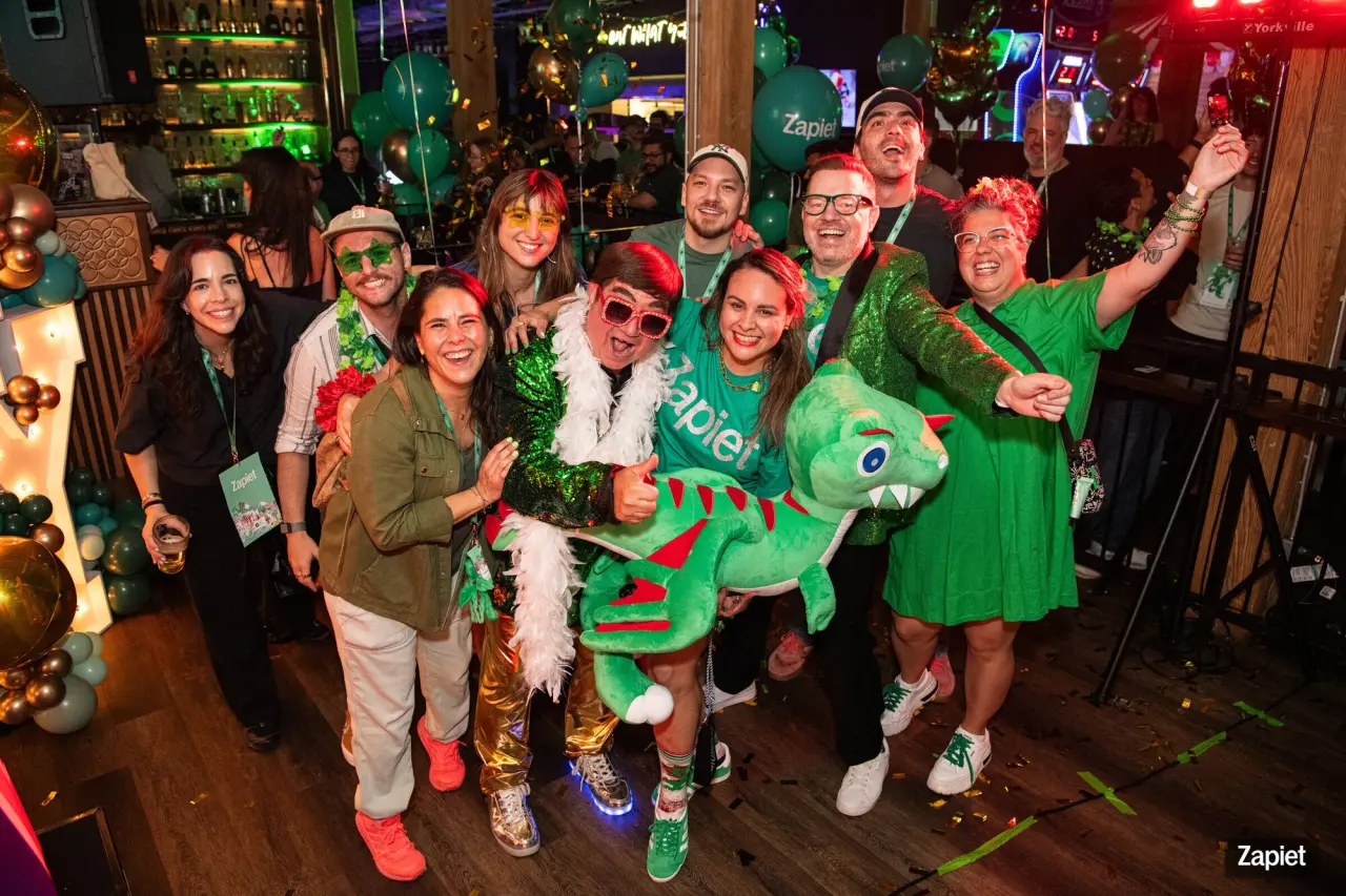 The Elton John tribute performer poses with the Zapietousaurus plush and a group of party guests in green outfits, surrounded by balloons and confetti at Zapiet's YOLO Party.