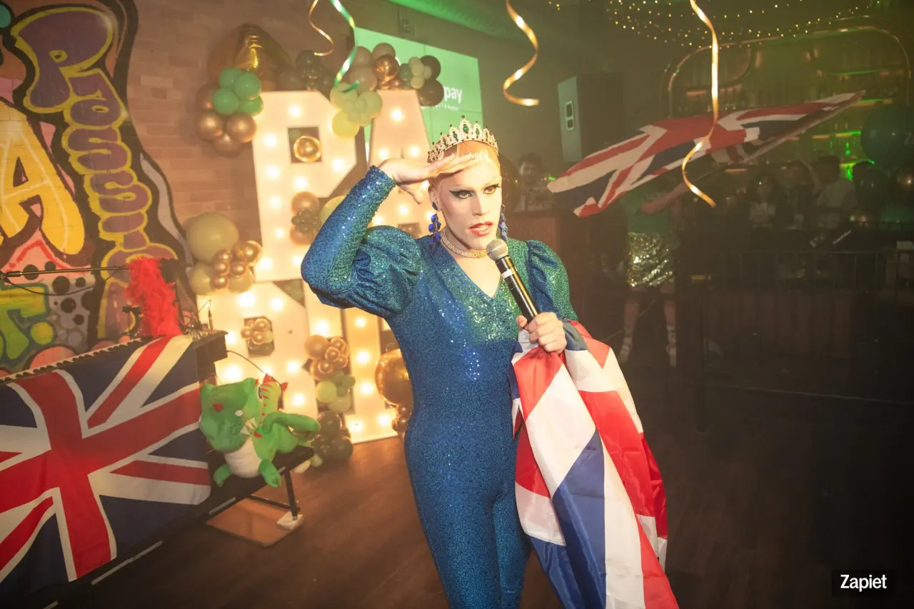 Drag queen Messy Margaret performs on stage in a sequined blue jumpsuit and tiara, holding a microphone and a Union Jack flag, in front of a lit-up marquee letter display at Zapiet's YOLO Party.