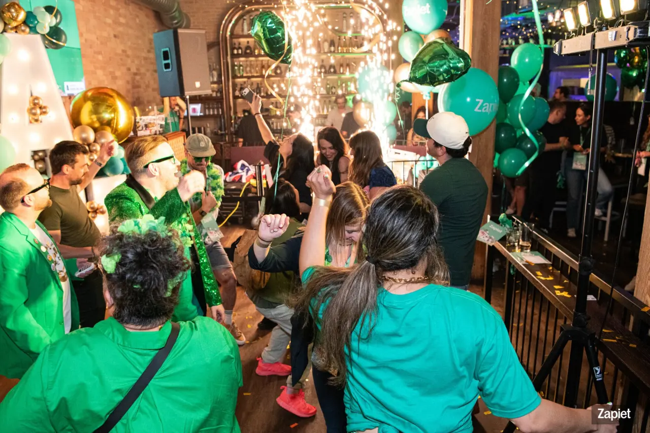 A crowd dressed in green dances and cheers at Zapiet's YOLO Party, with gold and green balloons, sparkler fireworks, and confetti filling the bar behind them.
