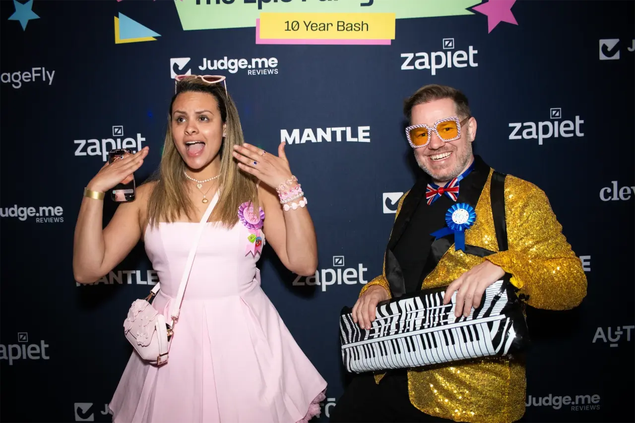 Zapiet's Emili Horncastle dressed as Barbie and Andrew Cargill dressed as Elton John, posing in front of the Epic Party's 10 Year Bash photo wall featuring sponsor logos including Judge.me, PageFly, Mantle, Cleverific, and AutoDS.